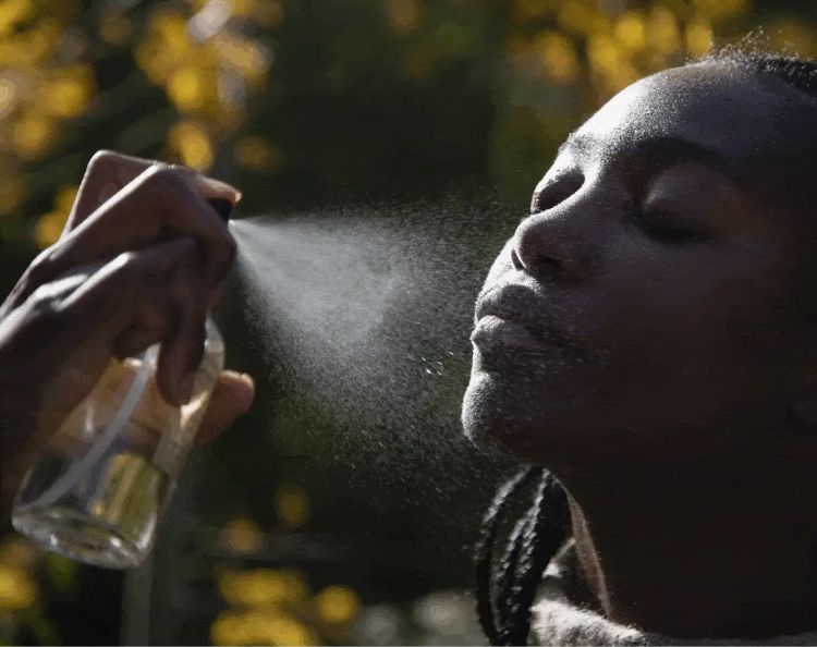 Person with eyes closed receiving a facial mist spray outdoors against a sunlit blurred background