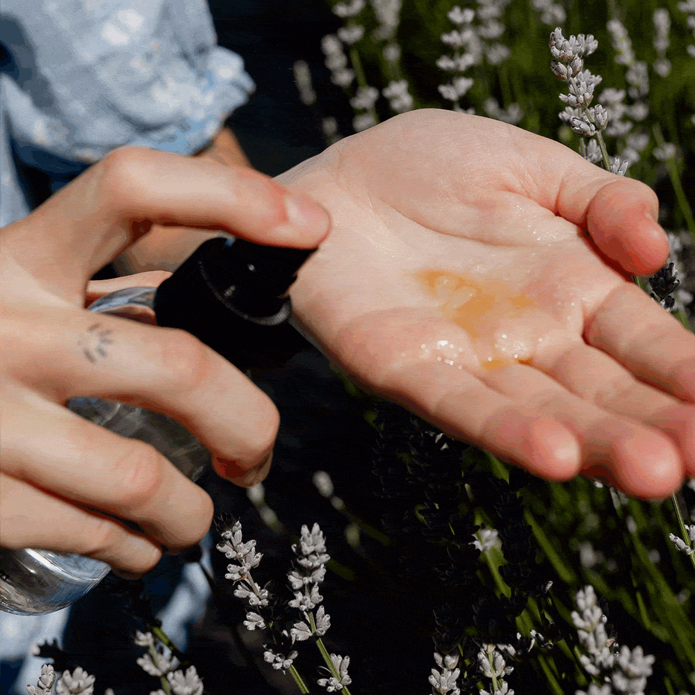 Hands demonstrate spraying a facial mist into an open palm over lavender flowers