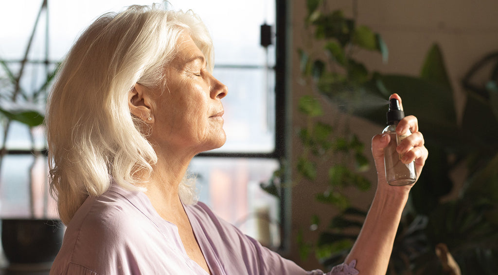 Woman with closed eyes spraying hydrosol mist toward her face in a sunlit room with plants in the background