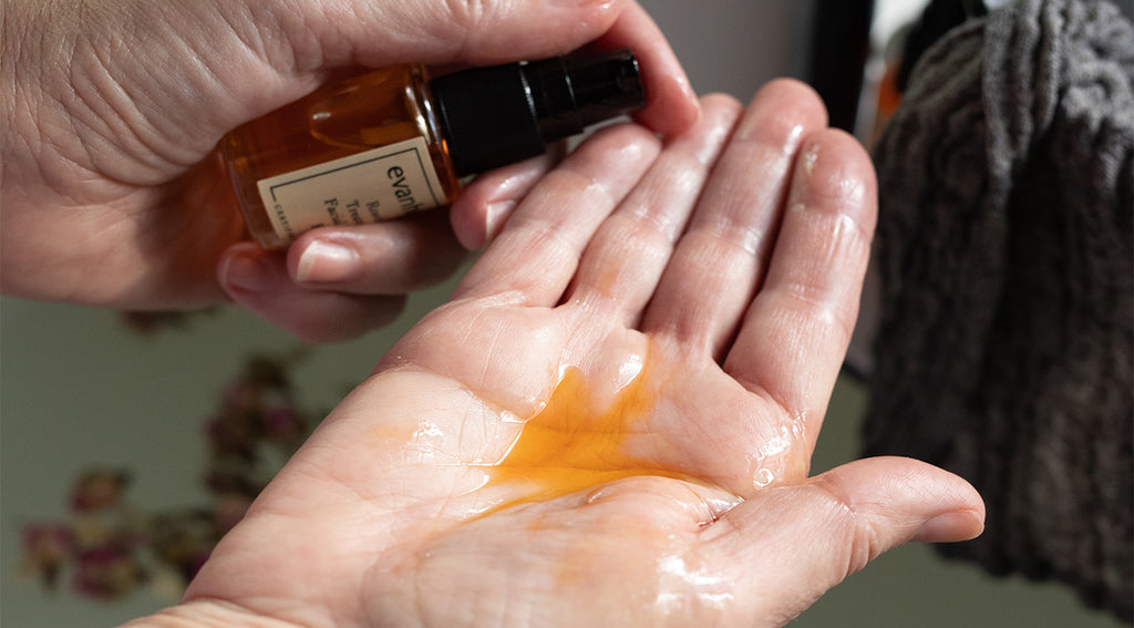Hands dispensing golden oil serum from an amber evanhealy bottle into palm for application