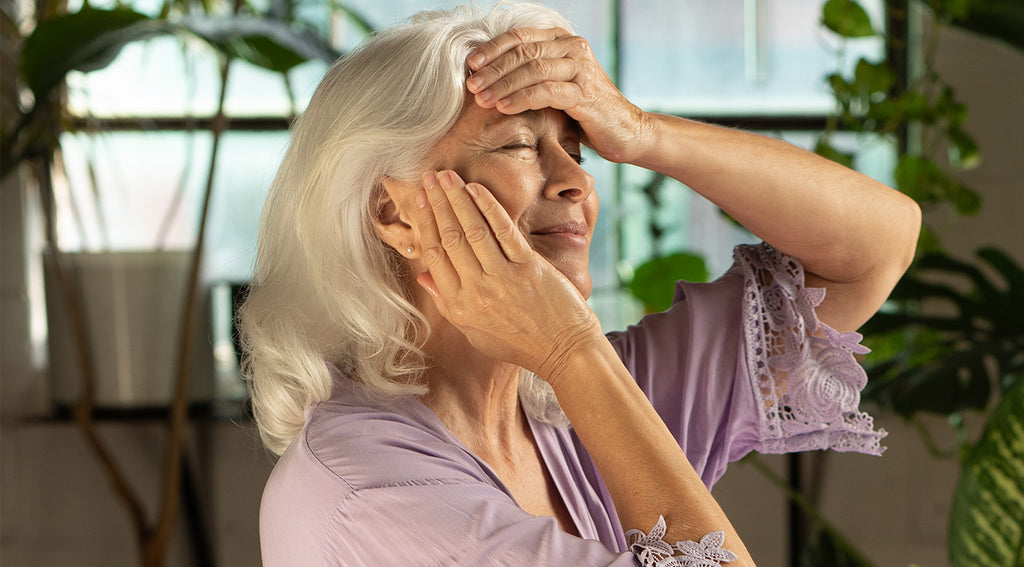 Woman pressing an oil-and-hydrosol mixture into her cheek during Step 5 of the ritual, wearing a lavender robe in a bright room with plants