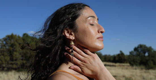 Woman with long dark hair touching her neck and jaw with eyes closed in a sunlit meadow under a clear blue sky