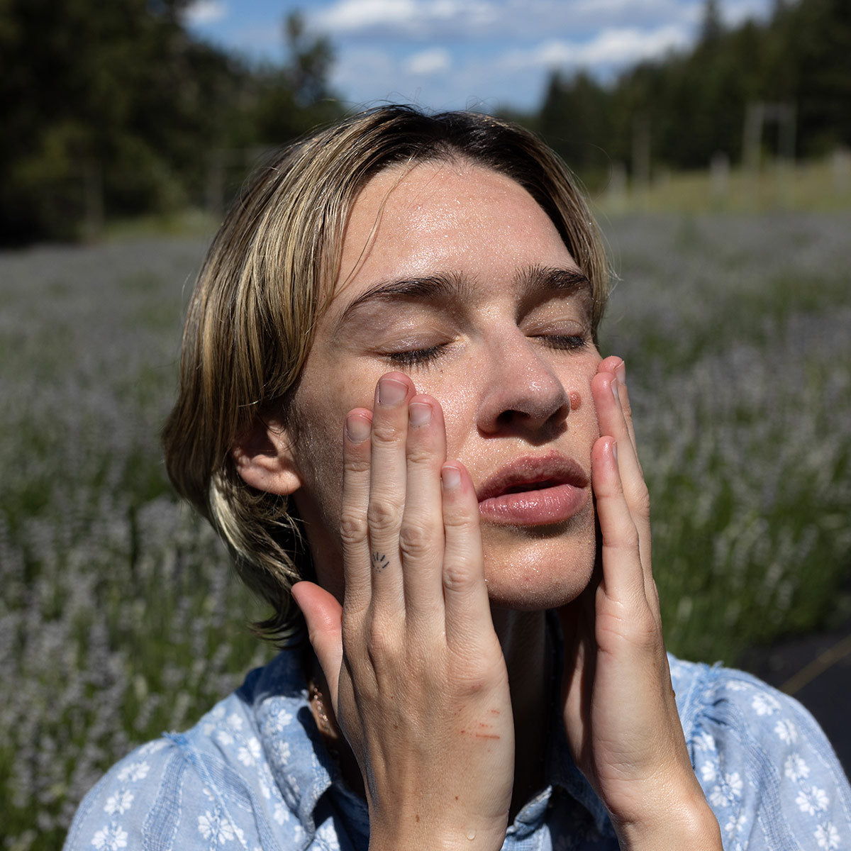 Person gently pressing hands to a damp face with eyes closed in a sunlit lavender field, wearing a light blue floral shirt