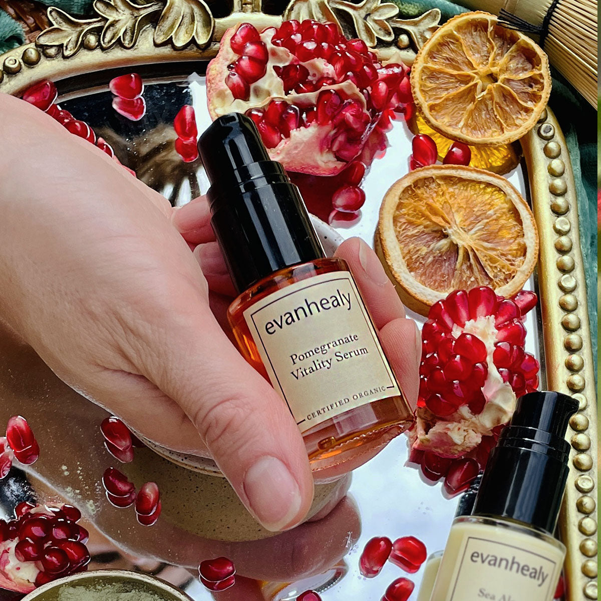 Close-up of Pomegranate Vitality Serum pump bottle held in a hand among pomegranates and dried citrus