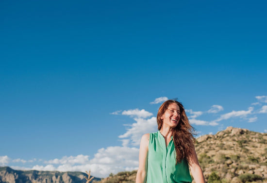 Smiling woman in a green sleeveless blouse with wind-blown hair standing outdoors against a blue sky and rocky hills