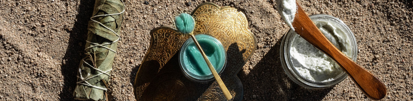 Two open jars of blue-green balm and white cream on desert sand with a sage bundle, brass tray, and wooden spatula