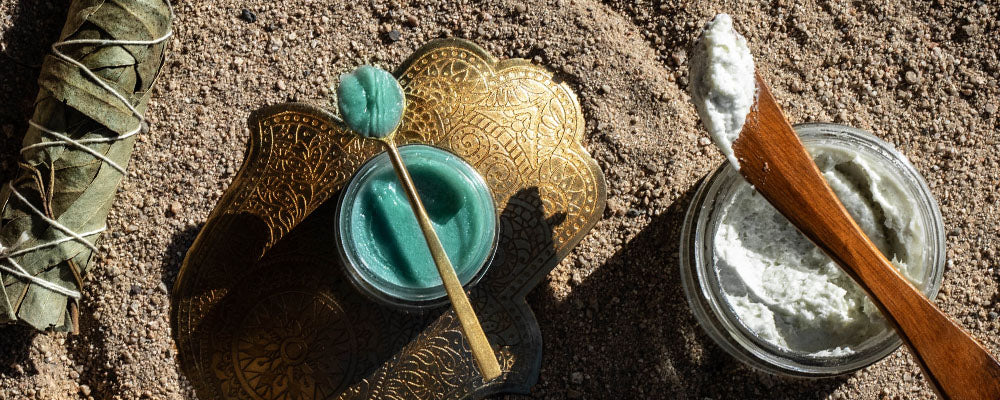 Two open jars of blue balm and white cream with wooden spatulas on sand beside a sage bundle