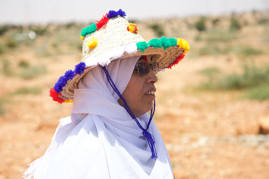Woman wearing a white headscarf and sunglasses with a straw hat decorated with colorful pom poms in a sunlit arid landscape