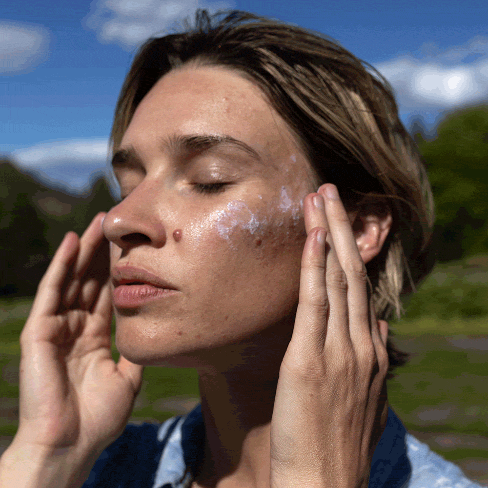Woman applying Sheer Mineral Sunscreen to her cheek outdoors with a blurred mountain background