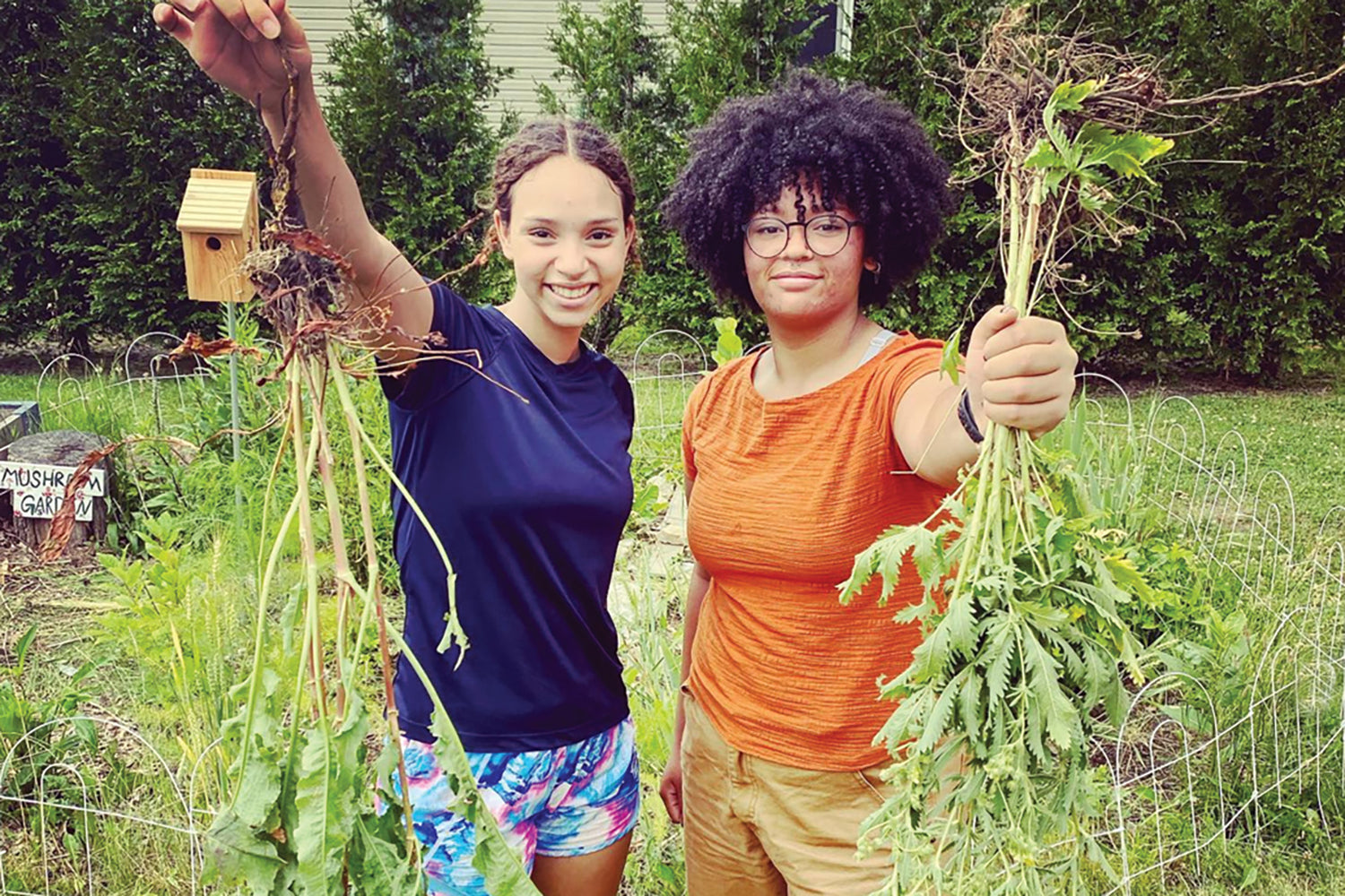 Two high school students holding freshly pulled herbs in a school garden, smiling in front of raised beds and shrubs