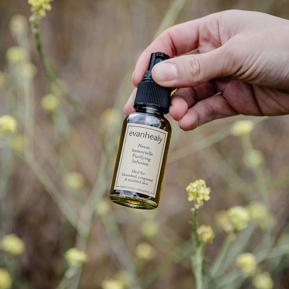 Close-up of Neem Immortelle Purifying Infusion bottle showcasing its dropper cap and label with flowering plants in background